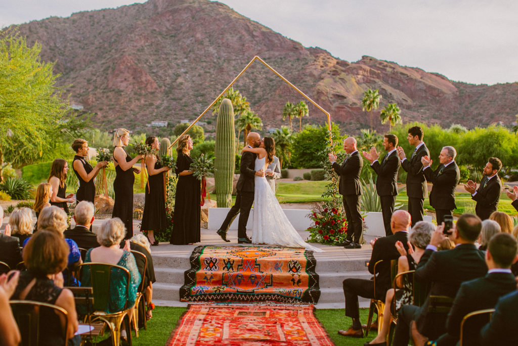 Paradise Valley Wedding - Geometric arch with greenery and red wedding flowers for the outside ceremony at this modern wedding and fall Arizona wedding. The greenery with warm colors wedding color palette was seen in the wedding flower arrangements, centerpieces, and arch flowers. Greenery bouquets, greenery boutonnières, and greenery with floral arrangements were also included. The fall wedding flower colors included a range of red, pink, orange, and peach, perfect to match the fall desert wedding sunset. Photos by Olivia Markle. #fallwedding #weddingbouquets #weddingflowers #weddings #weddingceremony