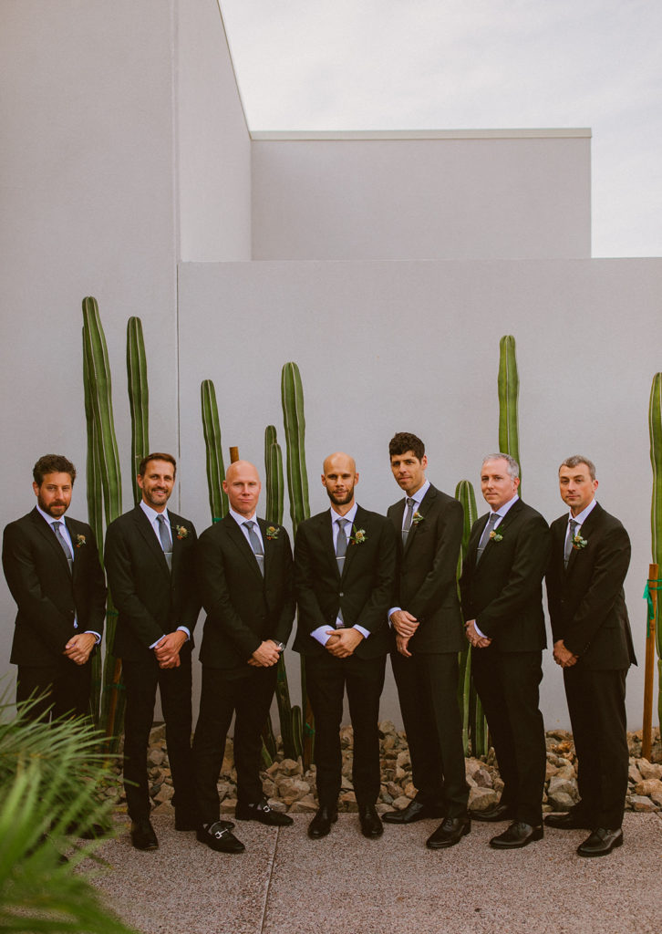 Paradise Valley Wedding - Groomsmen in black suits at Mountain Shadows modern wedding and fall Arizona wedding. The greenery with warm colors wedding color palette was seen in the wedding flower arrangements, centerpieces, and arch flowers. Greenery bouquets, greenery boutonnières, and greenery with floral arrangements were also included. The fall wedding flower colors included a range of red, pink, orange, and peach, perfect to match the fall desert wedding sunset. Photos by Olivia Markle. #fallwedding #weddingboutonnieres #weddingflowers #weddings