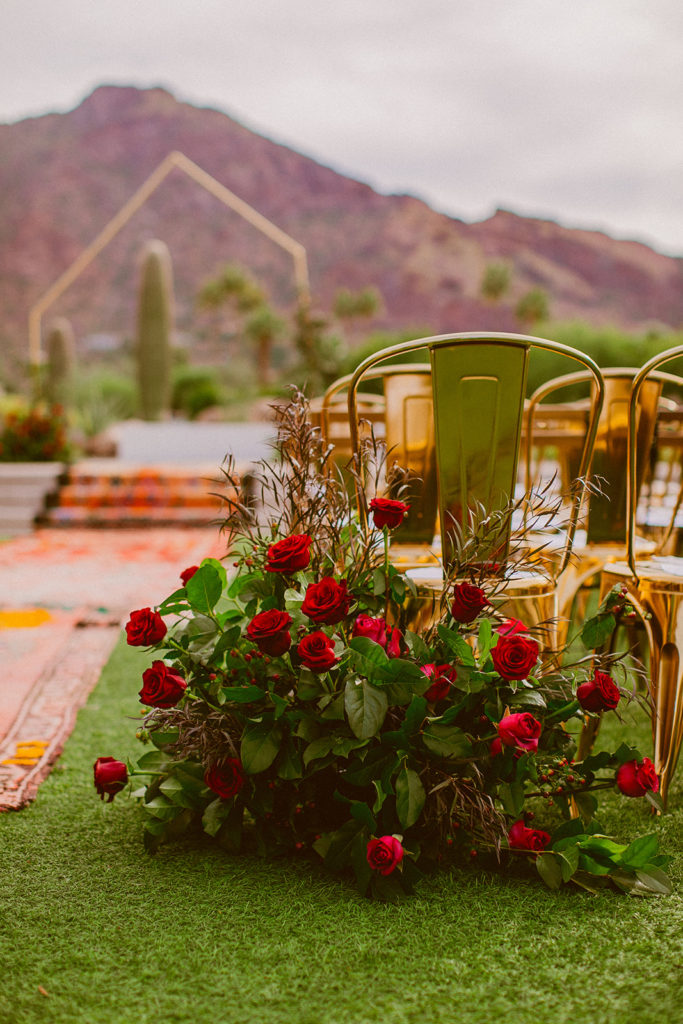 Paradise Valley Wedding - Red wedding flowers with greenery at aisle entrance for the outside ceremony at this modern wedding and fall Arizona wedding. The greenery with warm colors wedding color palette was seen in the wedding flower arrangements, centerpieces, and arch flowers. Greenery bouquets, greenery boutonnières, and greenery with floral arrangements were also included. The fall wedding flower colors included a range of red, pink, orange, and peach, perfect to match the fall desert wedding sunset. Photos by Olivia Markle. #fallwedding #weddingbouquets #weddingflowers #weddings #weddingceremony