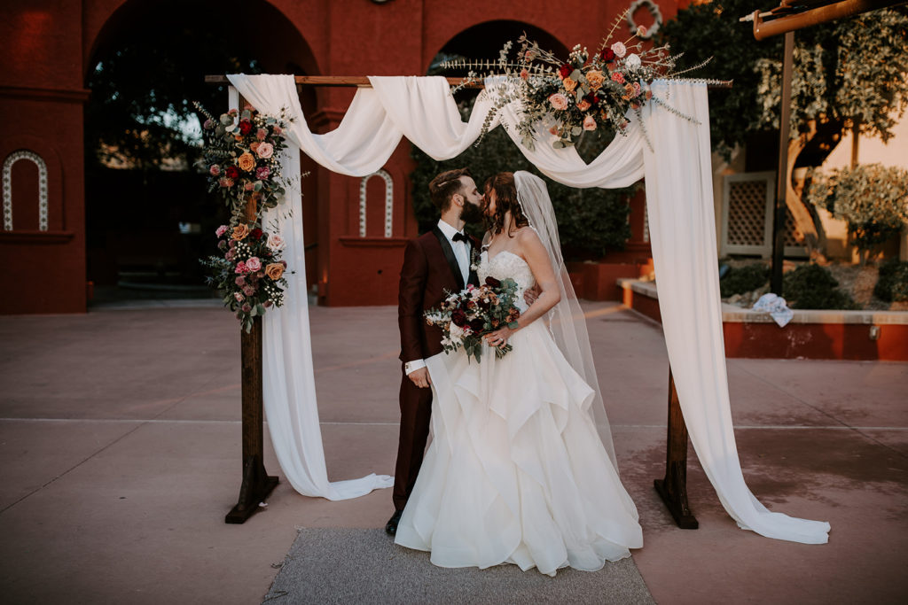 Bride and groom kissing in front of wedding arch with floral arrangements and white draped fabric.