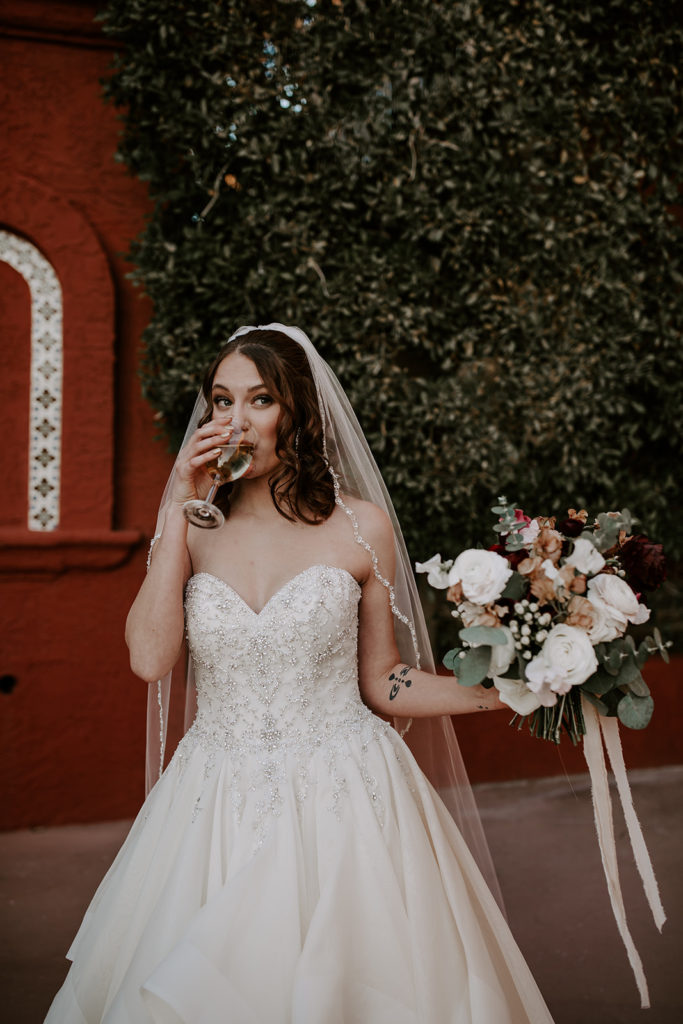 Bride holding bouquet and drinking out of a glass.