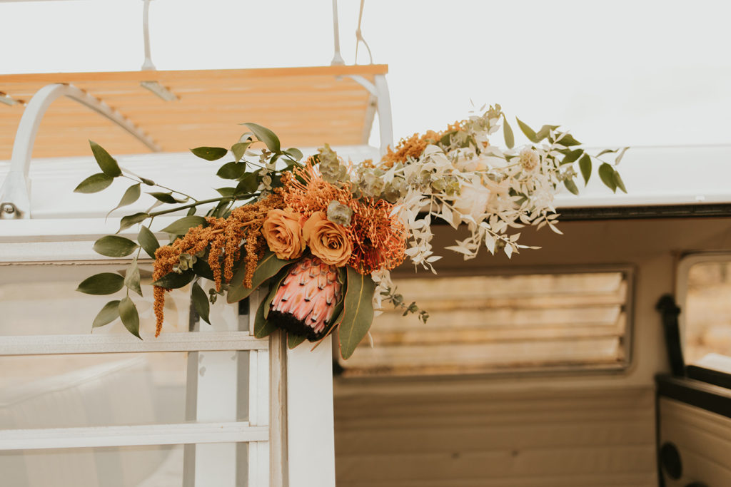 Bus floral accent arrangement with queen protea in pink, with orange and white and greenery.