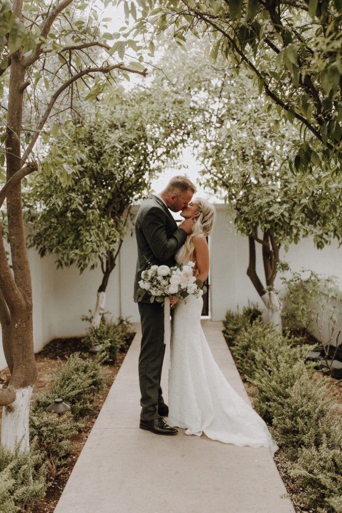 Bride and groom kissing on sidewalk lined with desert plants.