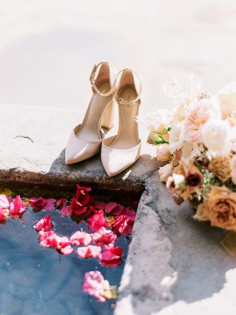 Bride's shoes and bouquet next to water pool.