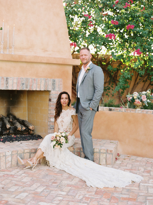 Bride sitting in front of outdoor fireplace, groom standing next to fireplace at Royal Palms.
