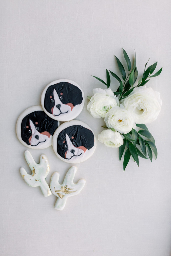 Cookies with dog image on them, and cactus cookies next to white flowers with greenery.