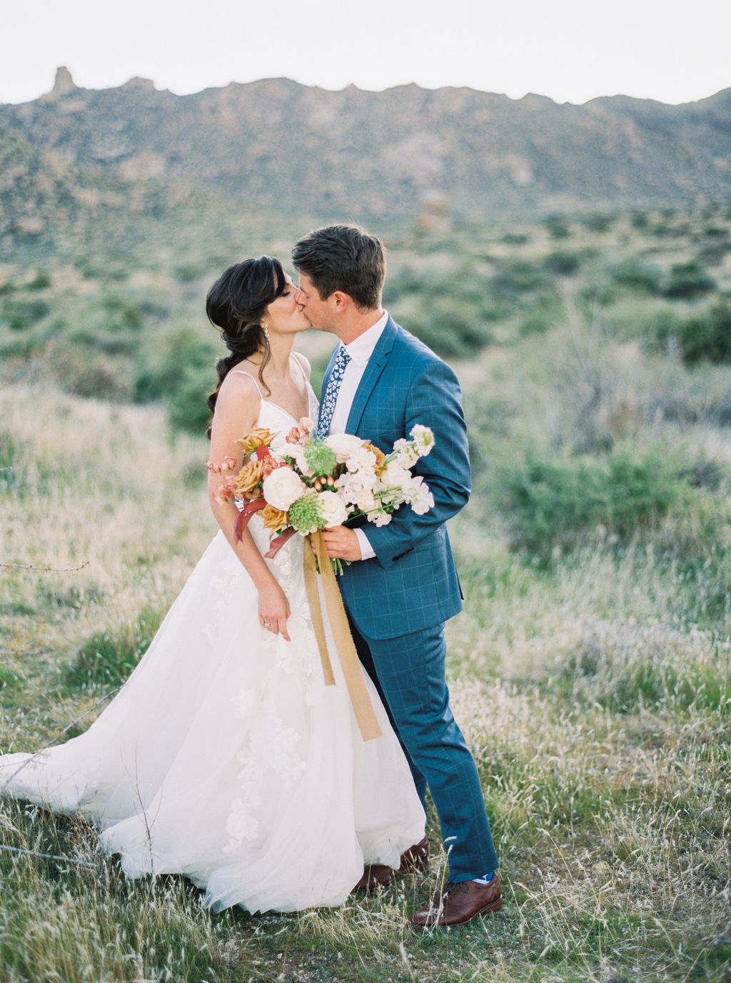 Bride and groom kissing in a field of wild grass with mountains in the back, groom holding a bouquet with trailing gold ribbon.
