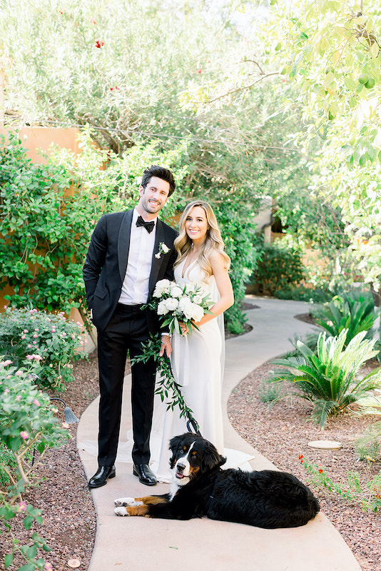 Bride and groom standing next to each other on outdoor sidewalk, holding dog leash with greenery for black medium sized dog.