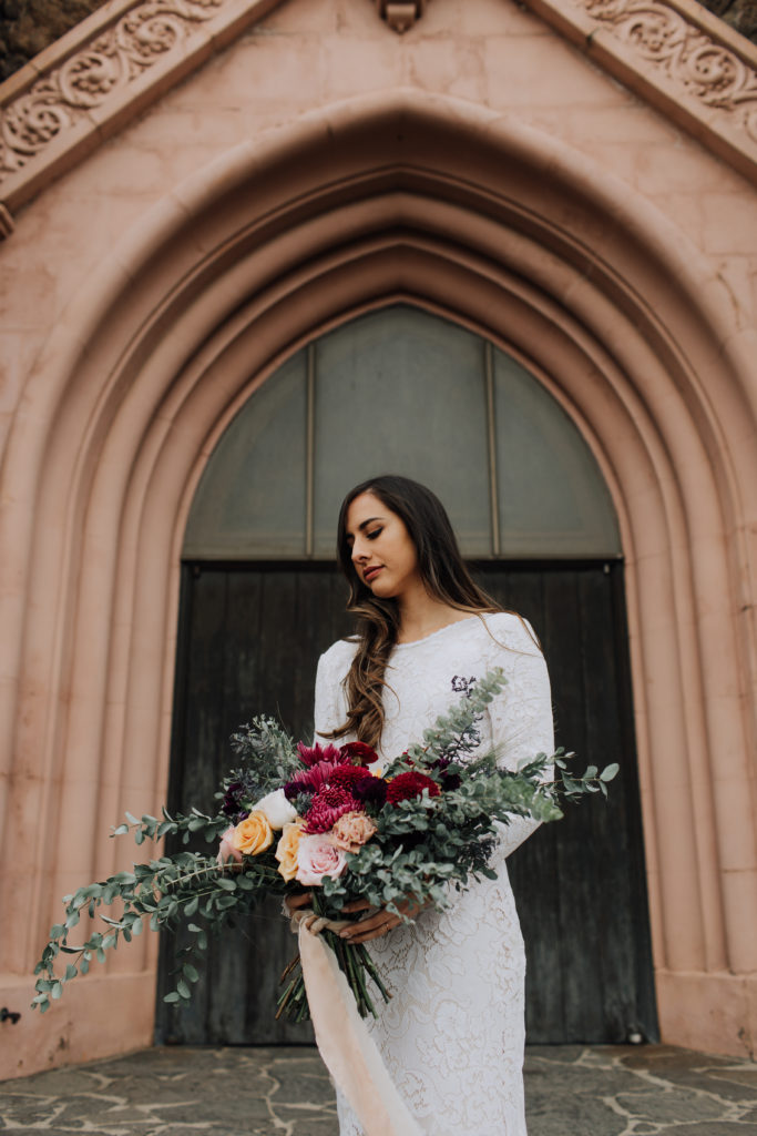 Bride standing in front of pink colored church looking down at bouquet she is holding.