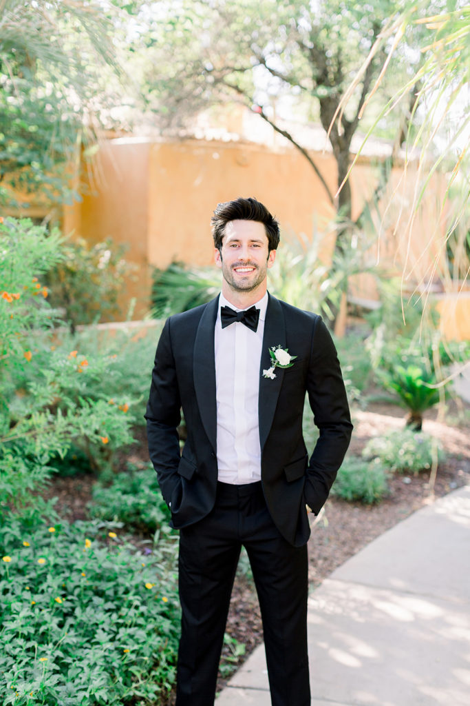 Groom smiling at camera, in black tux with black bowtie and white flower boutonniere.