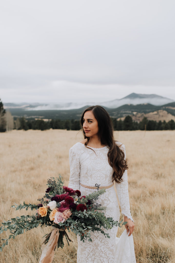 Bride holding bouquet of maroon, yellow, blush and white flowers with greenery standing in field of dried grass in front of mountain in distance in Flagstaff.