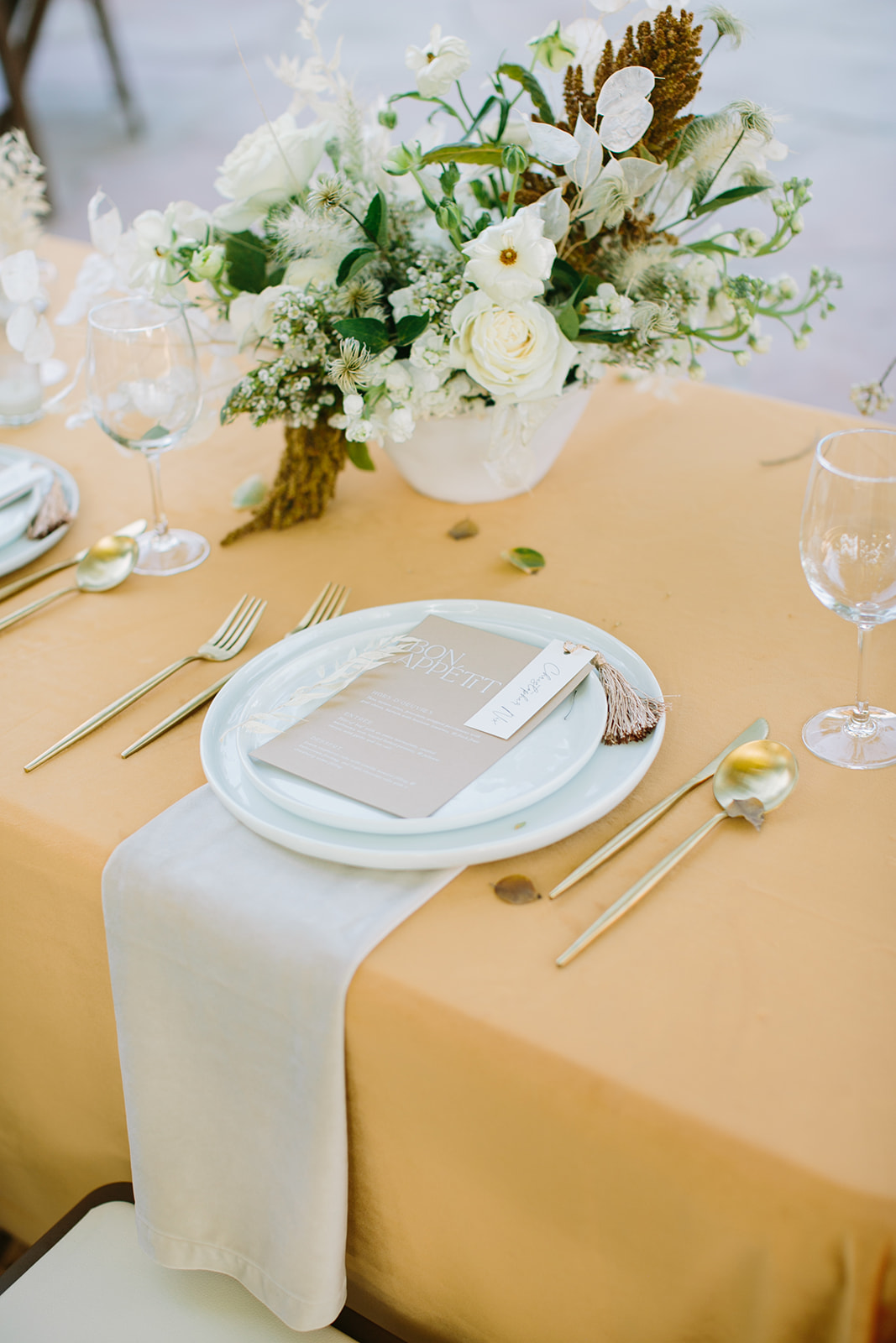 Wedding reception place setting with white plates, gold silverware and mustard linens.