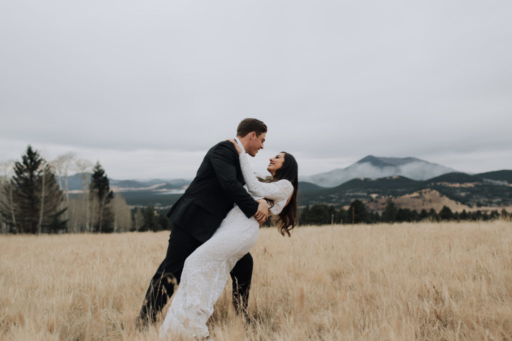 Groom dipping bride to side in field of dried grass in Flagstaff with mountain in distance.
