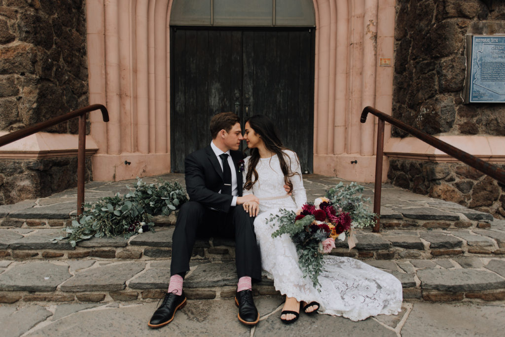 Bride and groom sitting on stone steps touching noses together, bride holding bouquet, with greenery behind them on ground.