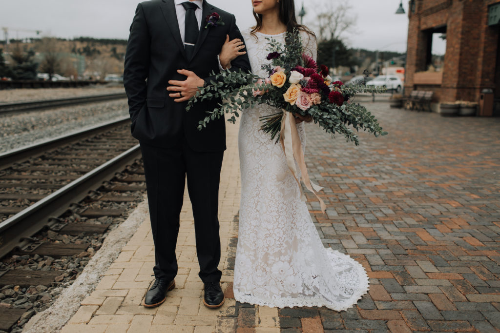 Bride with hand around groom's arm standing with bouquet at empty train depot by tracks.
