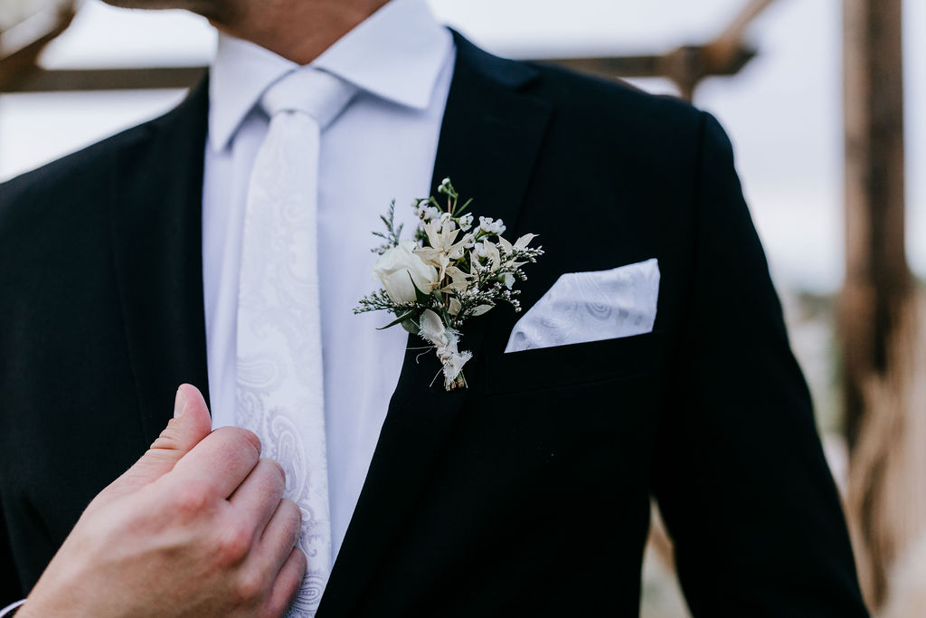White floral boutonniere with dried elements on groom with black suit and white paisley tie.
