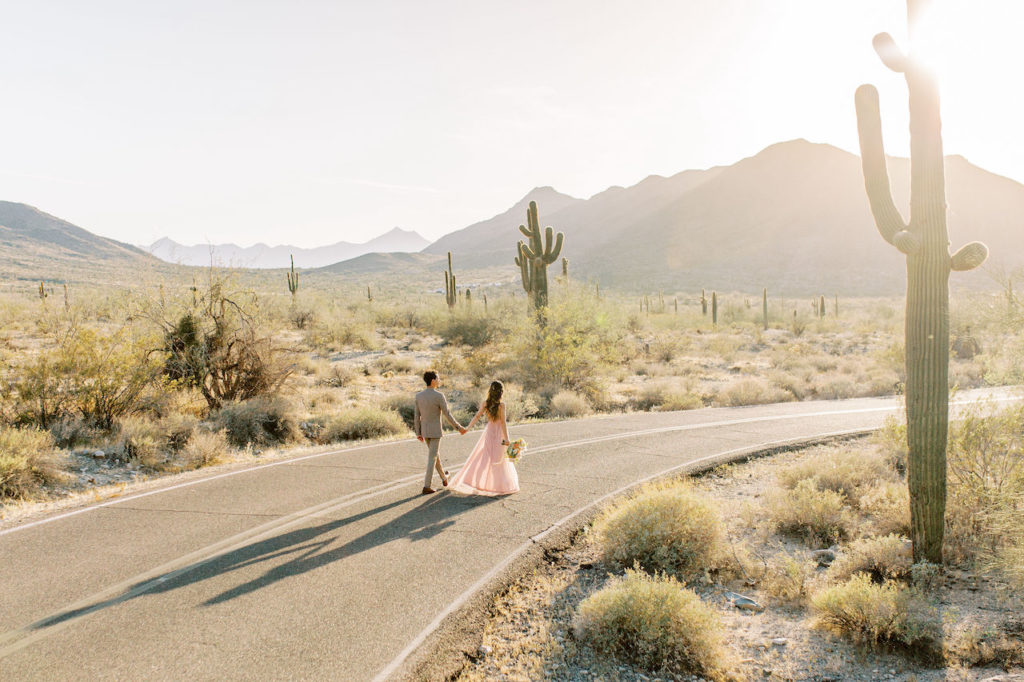 Husband and wife holding hands walking down two lane highway in middle of desert.