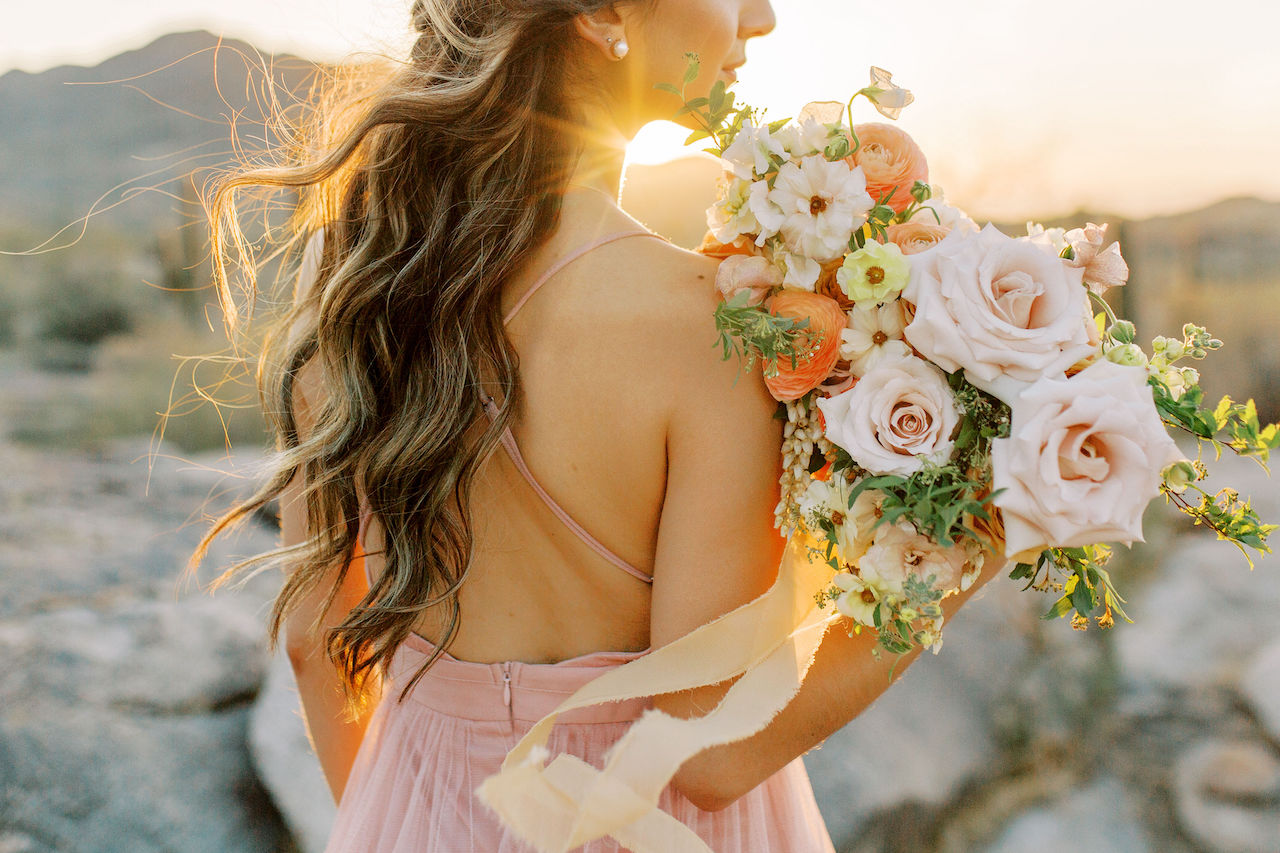 Back profile of woman in pink dress holding bouquet of lush flowers with sunsetting in distance.