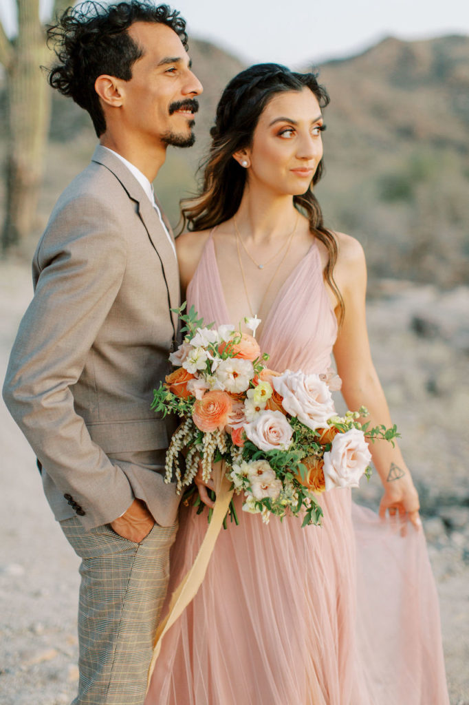 Husband and wife embracing, looking off while standing in desert. Woman holding lush bridal bouquet.