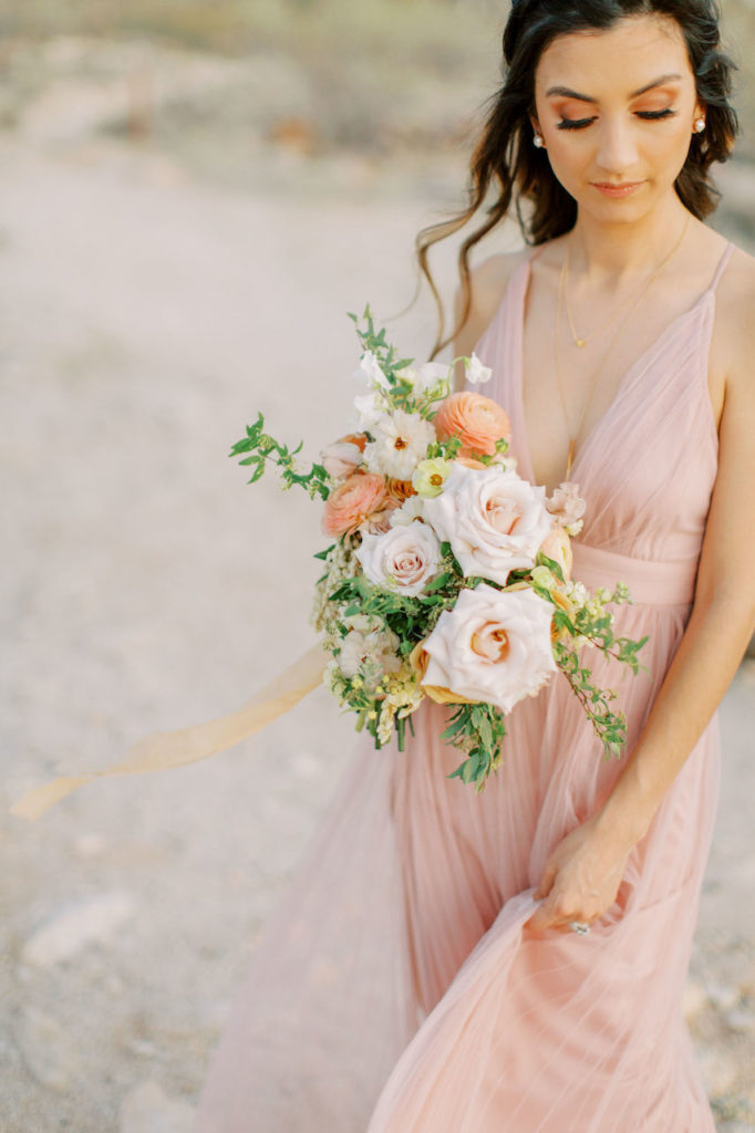 Woman in pink flowing dress holding bouquet of pink roses and peach ranunculus.