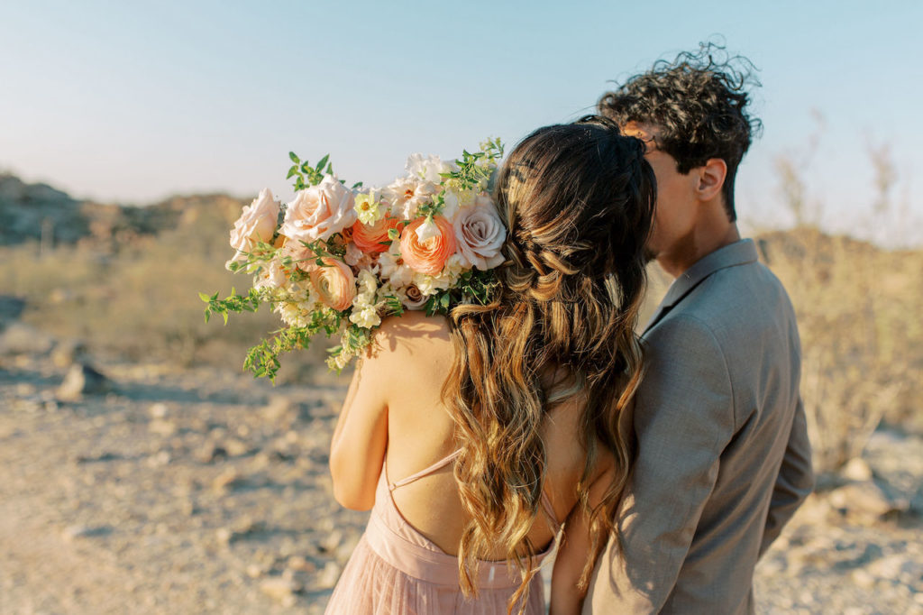 Back profile of wife and husband kissing while standing in desert and she is holding flower bouquet over her shoulder..