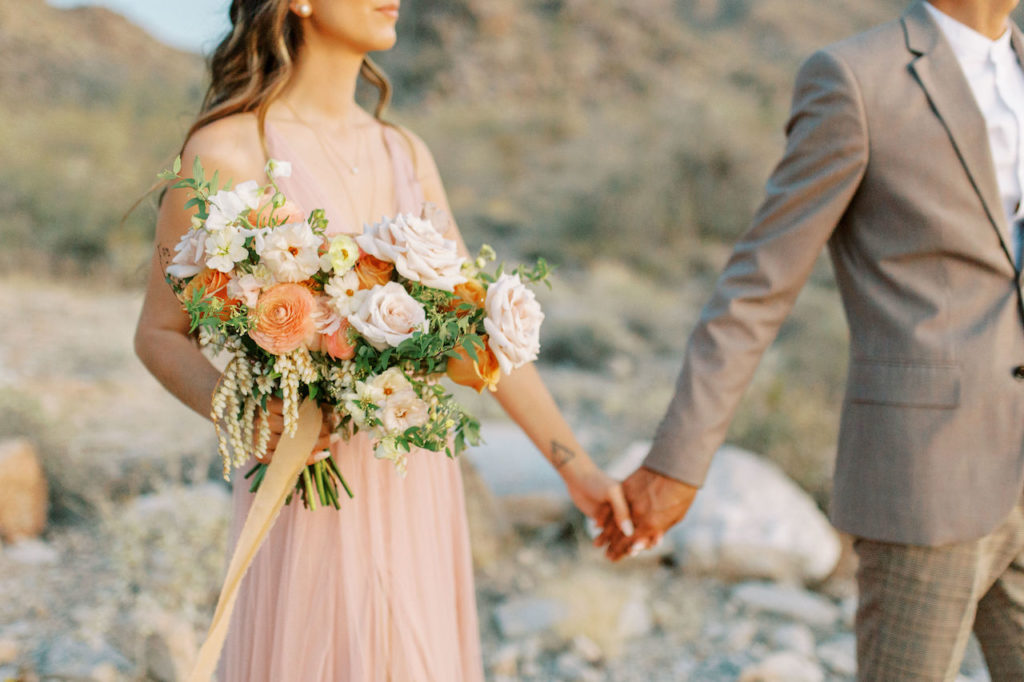 Woman holding bouquet and man's hand in tan suite standing in desert.