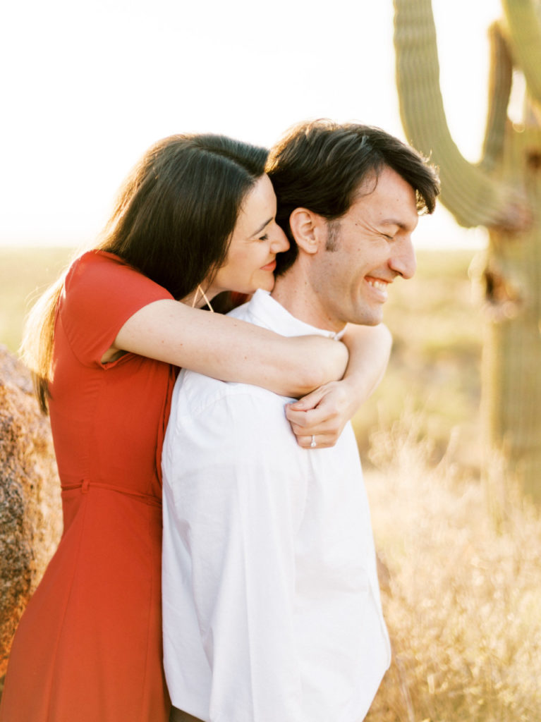 Woman hugging man from behind, both smiling, standing in desert.