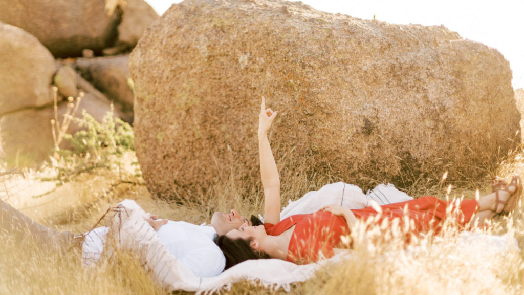 Couple laying down on blanked in front of boulder in desert, woman pointing up toward sky.