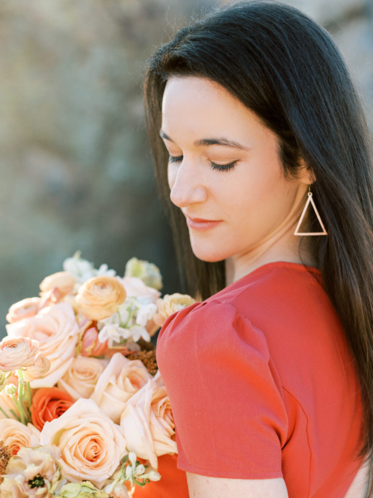 Woman in coral dress looking down her left side to ground, holding bouquet.