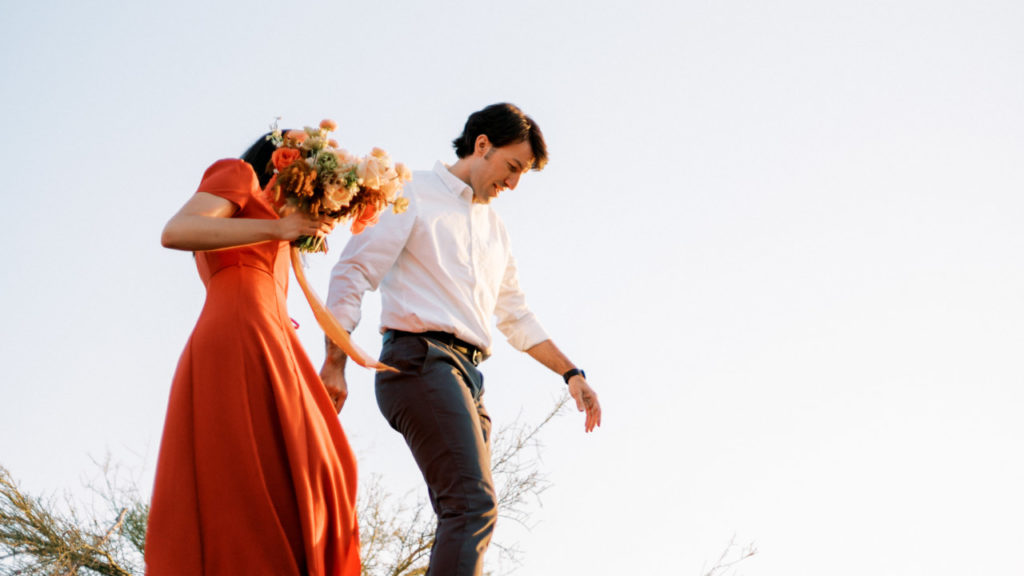 Woman in coral dress holding a bouquet and man in white shirt walking.