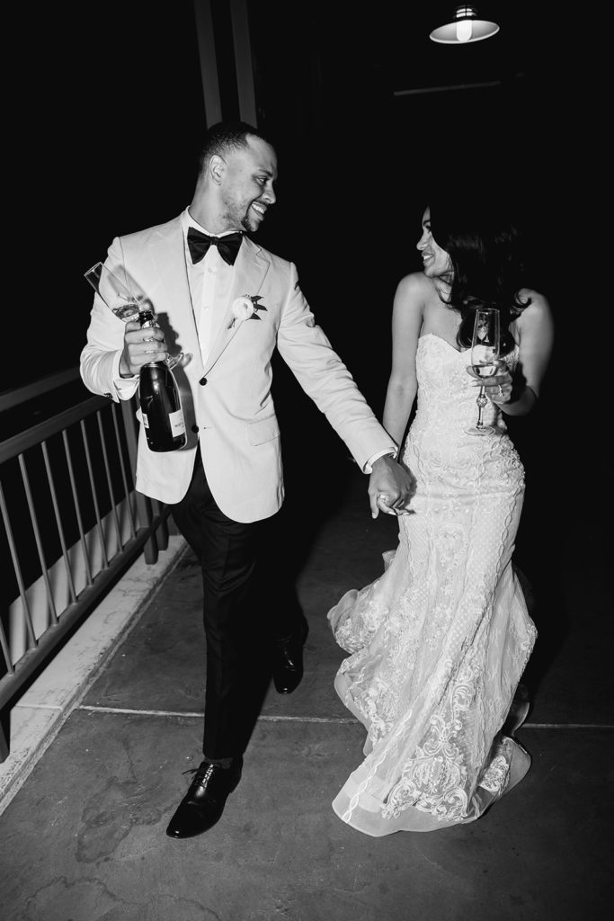 Bride and groom walking holding hands, groom holding champagne bottle.