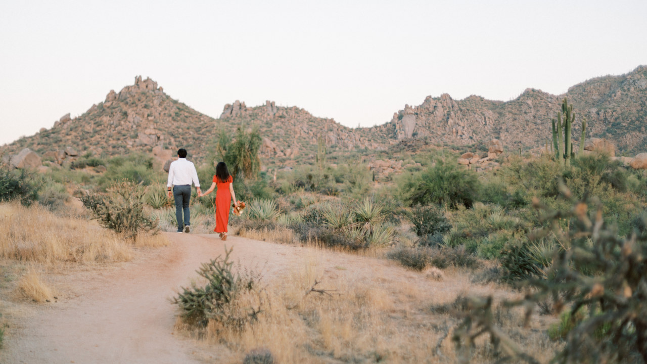 Woman in coral dress holding a bouquet and man in white shirt walking away in desert.