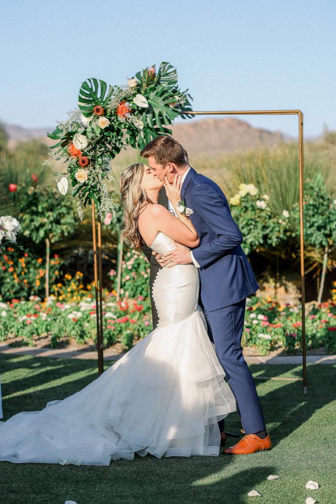 Bride and groom kissing in front of copper pipe arch with tropical themed floral arrangement.