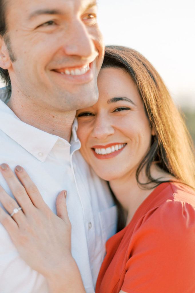 Woman smiling into camera, showing engagement ring on hand resting on chest of man looking off in white shirt.