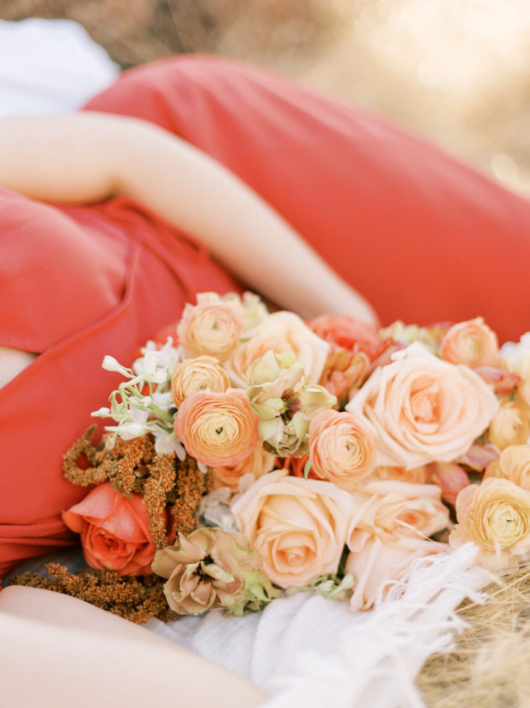 Woman in coral dress laying on white blanket with flower bouquet resting in front of her.
