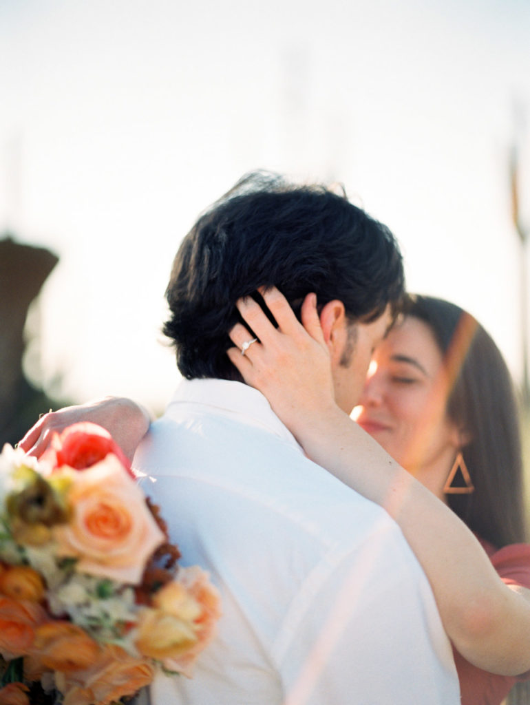 Woman putting hand on back of man's head while embracing, showing engagement ring, holding a bouquet.