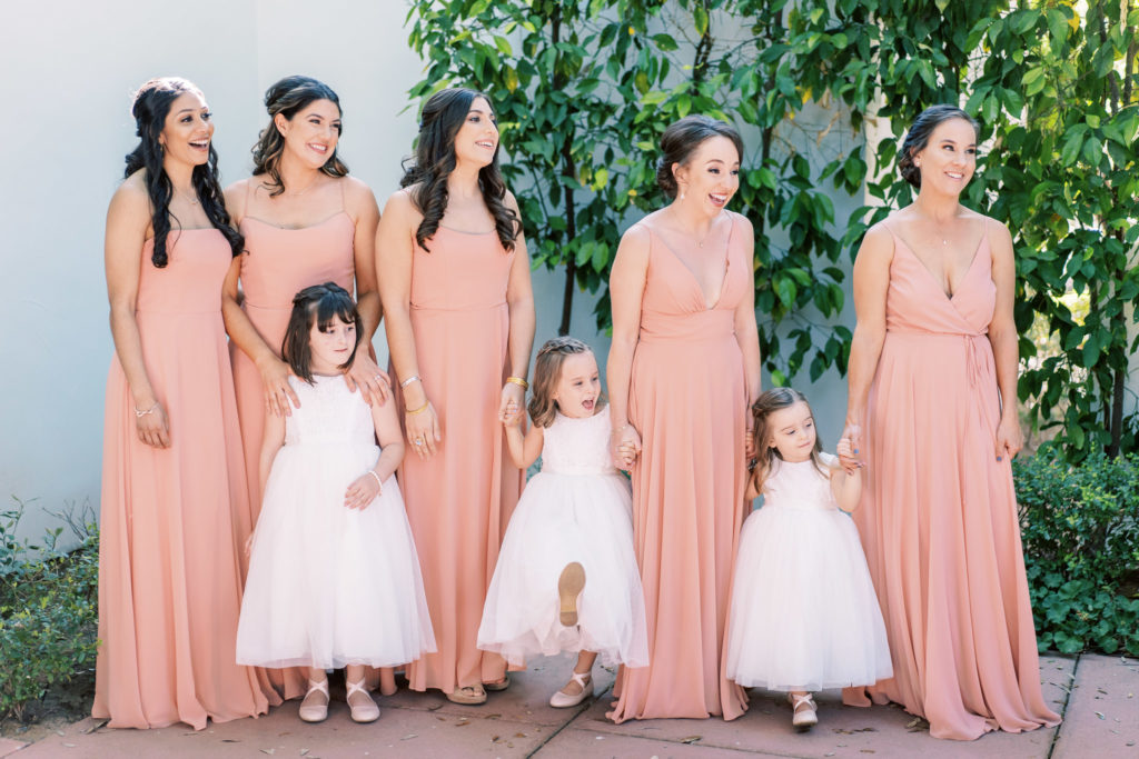 Bridesmaids in pink dresses holding hands with flower girls looking off.