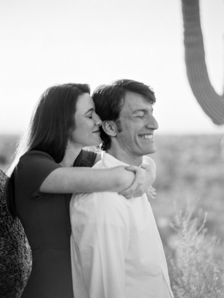 Woman hugging man from behind, both smiling, standing in desert.