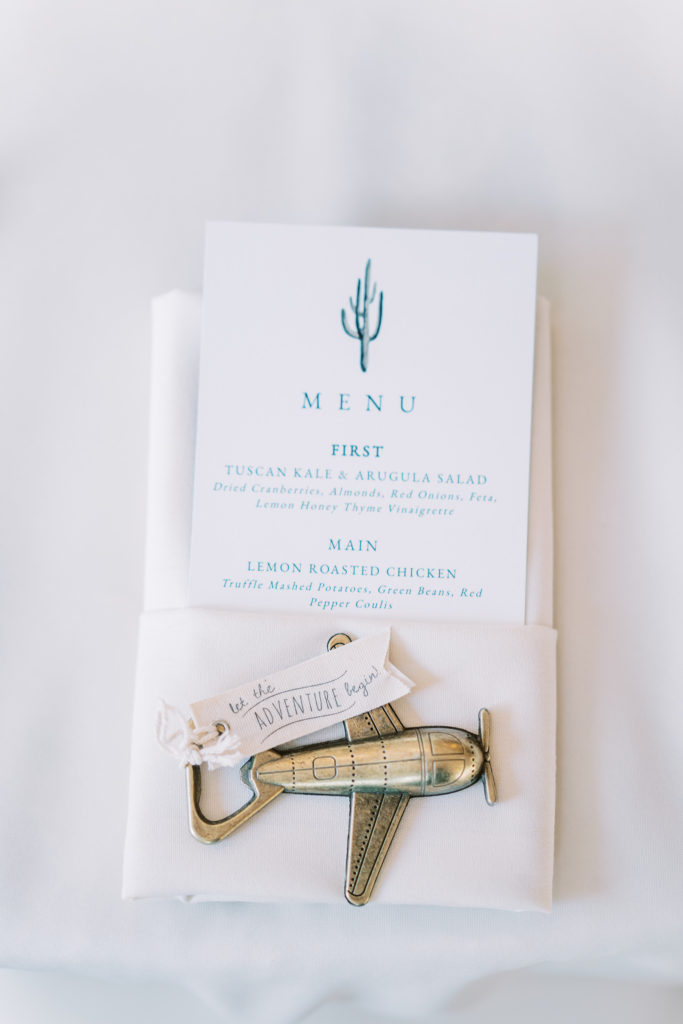 Wedding menu with cactus detail tucked into white napkin with guest gift of airplane bottle opener on top.