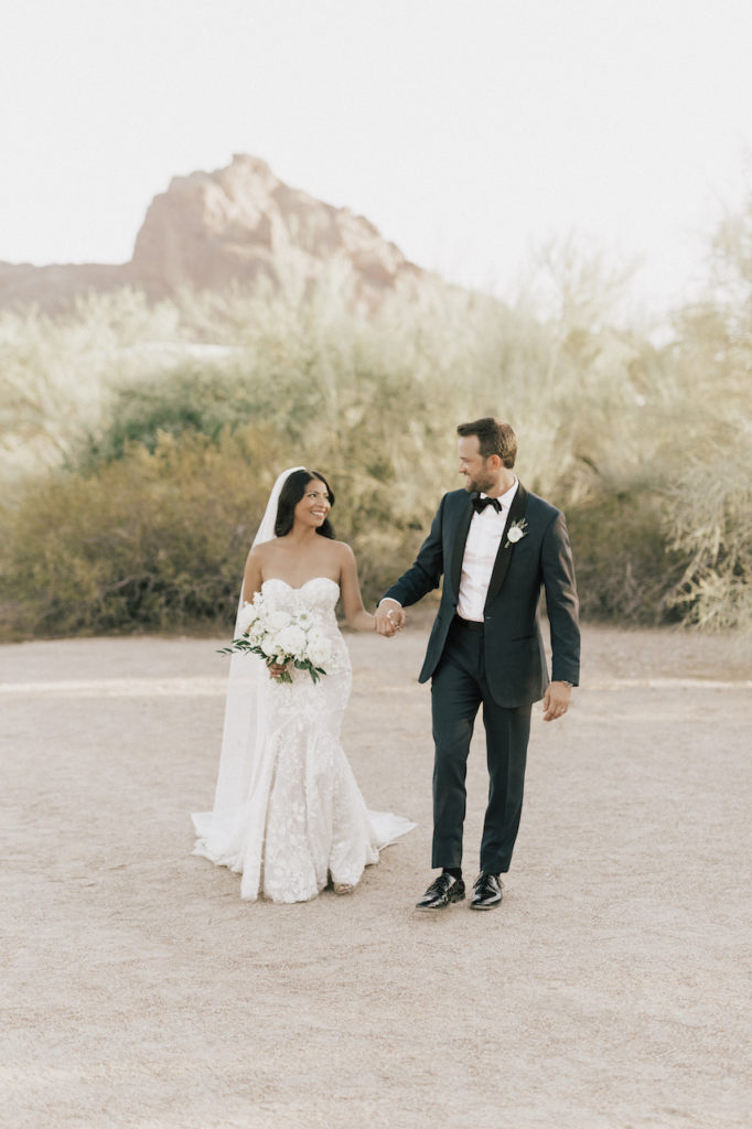 Bride and groom walking, holding hands, in desert landscape.