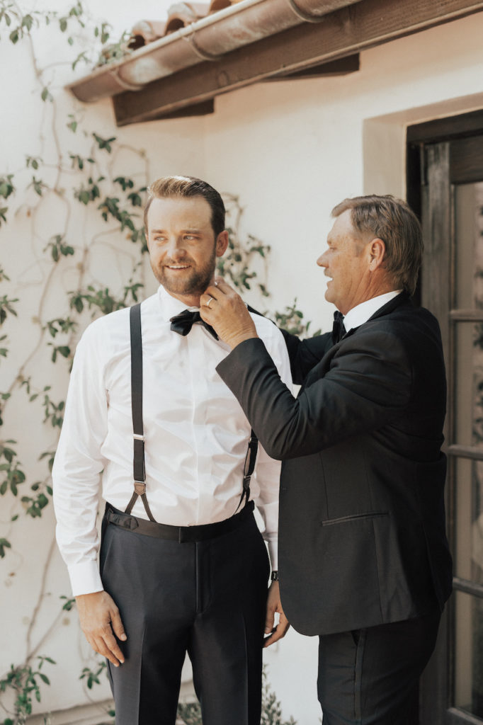 Groom receiving assistance from man putting on his bow tie.