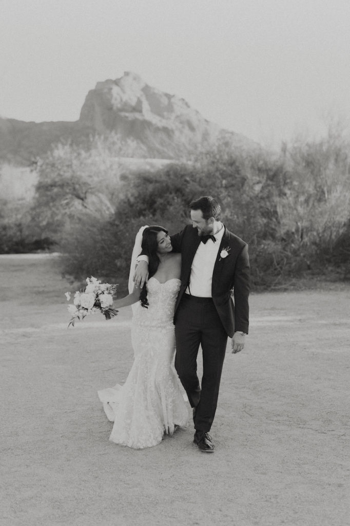 Bride and groom walking, smiling at each other, in desert landscape.