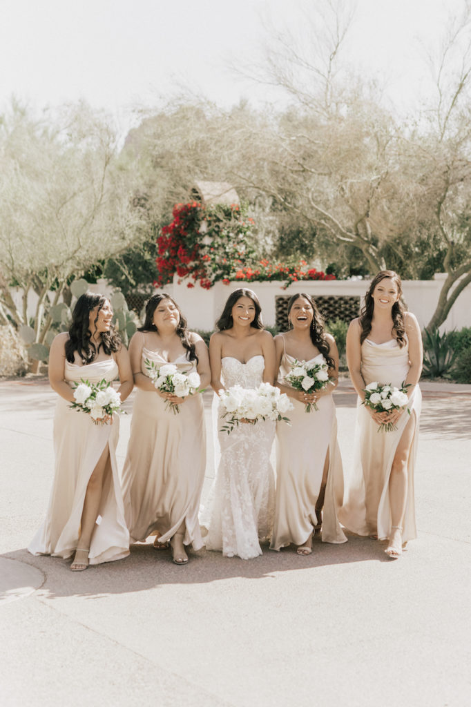 Bride walking with bridesmaids in taupe dresses, all holding bouquets and smiling.