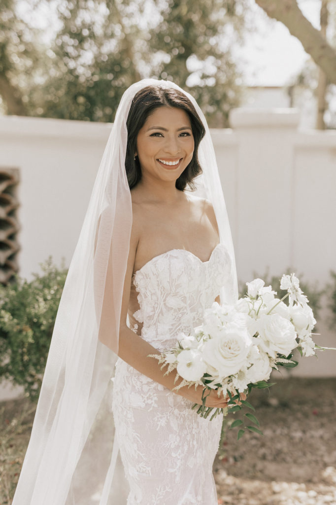 Bride smiling, wearing wedding dress and holding bouquet of lush white bridal bouquet.