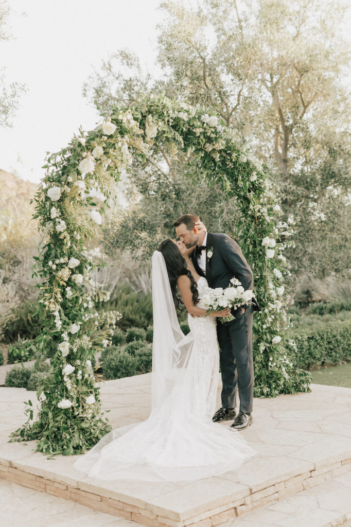 Bride holding white flowers bouquet kissing groom under floral and greens wedding arch at El Chorro.