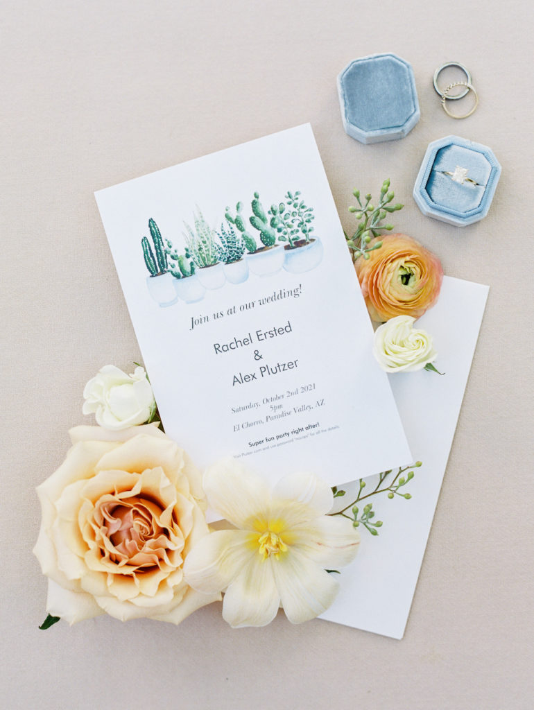 Cactus wedding invitation next to flowers and rings.