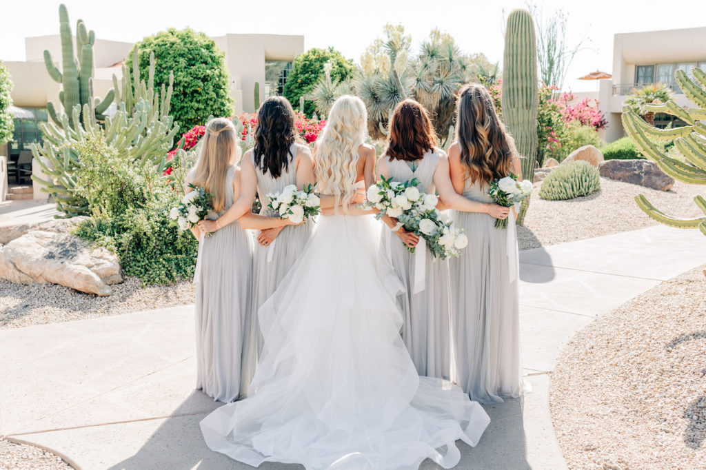 Bride and bridesmaids facing away, with arms around each other holding bouquets.