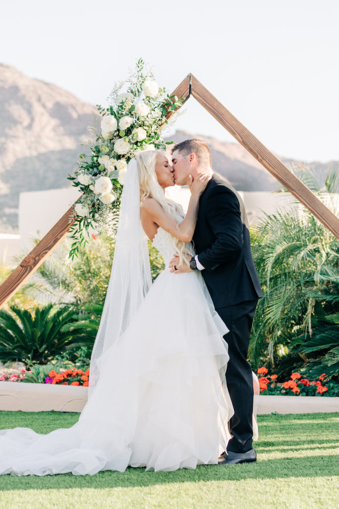Bride and groom kissing in front of wedding arch with Arizona Camelback mountains in background.