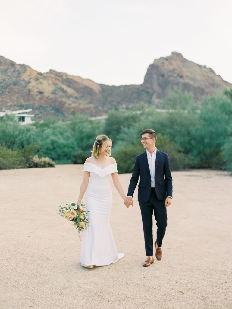 Bride and groom holding hands, walking in desert landscape.