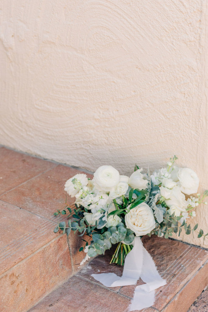 White flowers bridal bouquet with greenery sitting on tile step outside.